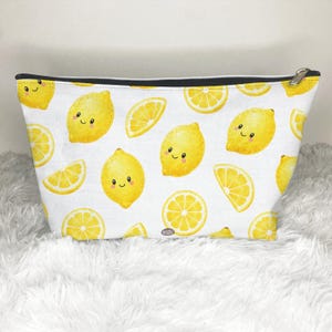 May include: A white zippered pouch featuring a repeating pattern of bright yellow lemons with smiling faces and yellow lemon slices. The pouch has a black zipper closure at the top and a textured fabric surface with a clear, colorful print.