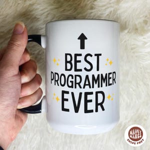May include: White ceramic mug with the text "BEST PROGRAMMER EVER" in black, with a black arrow and yellow star accents. The mug has a black handle and rim. A logo is visible in the bottom right corner.