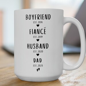 K&ouml;nnte beinhalten: Wei&szlig;e Keramiktasse mit schwarzem Text, der "Boyfriend Est. 2014", "Fianc&eacute; Est. 2019", "Husband Est. 2020" und "Dad Est. 2023" lautet.
