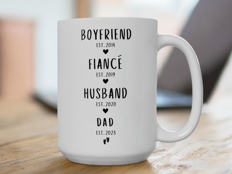 K&ouml;nnte beinhalten: Wei&szlig;e Keramiktasse mit schwarzem Text, der "Boyfriend Est. 2014", "Fianc&eacute; Est. 2019", "Husband Est. 2020" und "Dad Est. 2023" lautet.