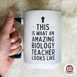 May include: White ceramic mug with a black handle and rim. The mug features the text "THIS IS WHAT AN AMAZING BIOLOGY TEACHER LOOKS LIKE" in black, with an upward-pointing arrow and star accents.