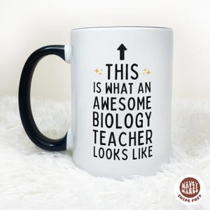May include: White ceramic mug with a black handle and rim. The mug features the text "THIS IS WHAT AN AWESOME BIOLOGY TEACHER LOOKS LIKE" in black, with an upward arrow and star accents.