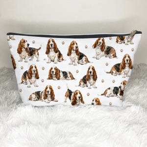 May include: White canvas cosmetic bag with a repeating pattern of Basset Hound illustrations and paw prints. The bag has a black zip and a gold-coloured zip pull. The bag is on a white, fluffy surface.