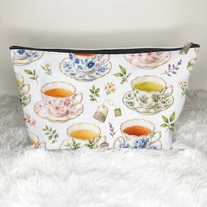 May include: A white zippered pouch featuring a repeating pattern of various teacups on saucers, tea bags, and small floral sprigs. The teacups are illustrated in different colors including blue, pink, and green, with delicate floral details.