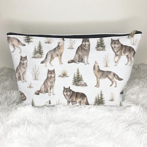 Pode incluir: Uma bolsa branca com fecho de correr apresentando um padrão repetitivo de lobos cinzentos em várias poses, incluindo sentados, de pé e a uivar, intercalados com pequenos pinheiros verdes e arbustos castanhos sobre um fundo claro.