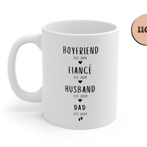 K&ouml;nnte beinhalten: Wei&szlig;e Keramiktasse mit schwarzem Text, der "Boyfriend Est. 2014", "Fianc&eacute; Est. 2019", "Husband Est. 2020" und "Dad Est. 2023" lautet.