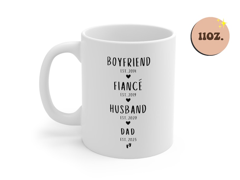 K&ouml;nnte beinhalten: Wei&szlig;e Keramiktasse mit schwarzem Text, der "Boyfriend Est. 2014", "Fianc&eacute; Est. 2019", "Husband Est. 2020" und "Dad Est. 2023" lautet.