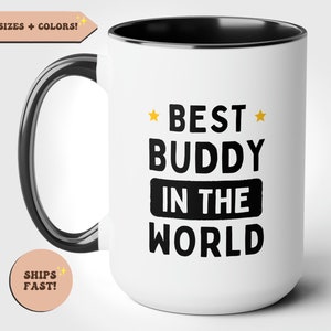 Puede incluir: Taza de cerámica blanca con borde y asa negros. La taza tiene texto negro que dice "*BEST* BUDDY IN THE WORLD" con dos pequeñas estrellas doradas.