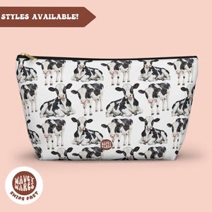 Op de afbeelding: Witte toilettas met rits, zwarte rand en een herhalend patroon van zwart-witte koeien. De tas is gemaakt van een zachte, duurzame stof.
