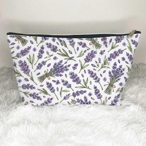 May include: A white zippered pouch featuring a repeating pattern of purple lavender sprigs and green stems. The design includes individual lavender flowers and small tied bundles of lavender scattered across the entire surface of the bag.