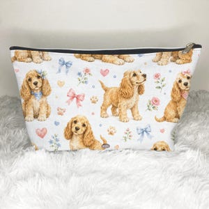May include: A white zippered pouch featuring a repeating pattern of golden-brown spaniel puppies, light blue and pink bows, small pink hearts, and floral accents. The puppies are depicted in various poses, including sitting and standing, with detailed fur.