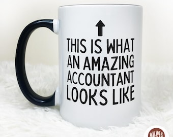 Könnte beinhalten: Weißer Keramikbecher mit schwarzem Henkel und Rand. Der Becher zeigt den Text "THIS IS WHAT AN AMAZING ACCOUNTANT LOOKS LIKE" in Schwarz, mit einem nach oben zeigenden Pfeil.