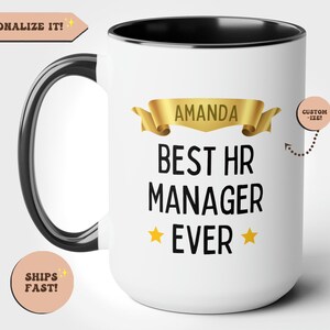 May include: White ceramic coffee mug with a black handle and rim. The mug features a gold ribbon with the text "AMANDA BEST HR MANAGER EVER" and two gold stars.
