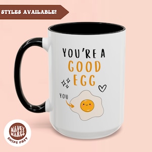 Może przedstawiać: Biały kubek ceramiczny z czarnym uchwytem. Kubek ma czarną obwódkę i przedstawia rysunkowy jajko sadzone z uśmiechniętą buzią i tekstem "You're a good egg" z sercem i błyskotkami.