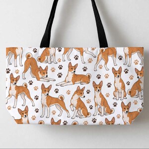 May include: A white tote bag with black shoulder straps featuring a repeating pattern of brown and white Basenji dogs in various poses, interspersed with small brown and black paw prints across the entire surface of the bag.