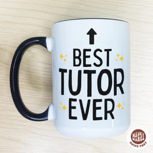 Op de afbeelding: Witte keramische mok met een zwart handvat en rand. De mok heeft de tekst "BEST TUTOR EVER" in vette zwarte letters, met een pijl en gele sterretjes. Inhoud ongeveer 325 ml.