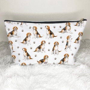 May include: White cosmetic bag with a repeating pattern of beagle dogs and paw prints. The bag has a black zip and a silver zip pull. The bag is on a white, fluffy surface. The beagle dogs are in a watercolor style.