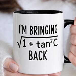 May include: White ceramic mug with a black interior and handle. The mug features the text "I'M BRINGING √(1 + tan²C) BACK" in black, a humorous reference to a mathematical formula. The mug is held by a person.