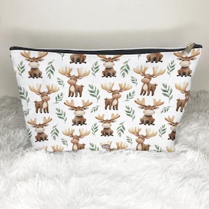 May include: White cosmetic bag with a repeating pattern of cartoon moose and green leaves. The moose are brown with large antlers. The bag has a black zipper and a gold-colored zipper pull.