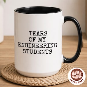 May include: White ceramic mug with a black interior and handle. The mug has the text "TEARS OF MY ENGINEERING STUDENTS" in black. The mug is on a woven coaster.