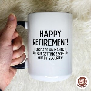 K&ouml;nnte beinhalten: Wei&szlig;er Keramikbecher mit schwarzem Henkel und Rand. Der Becher tr&auml;gt den Text "HAPPY RETIREMENT!" und eine humorvolle Botschaft &uuml;ber den Ruhestand. Eine Hand h&auml;lt den Becher.