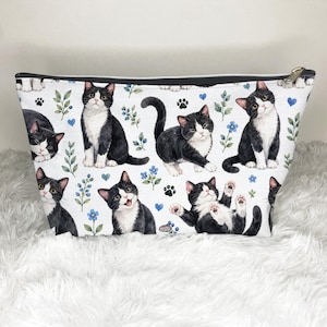 May include: A white zippered pouch featuring a repeating pattern of black and white tuxedo cats in various poses, interspersed with small green leafy sprigs, blue flowers, tiny blue hearts, and black paw prints against a clean white background.