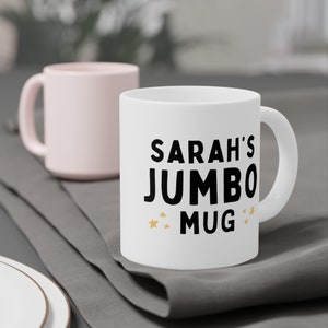 Może przedstawiać: Biały kubek ceramiczny z czarnym tekstem, kt&oacute;ry brzmi "Sarah's Jumbo Mug" ze złotymi gwiazdkami po obu stronach tekstu.