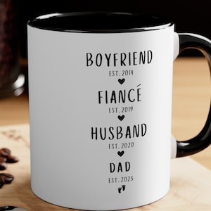 K&ouml;nnte beinhalten: Wei&szlig;e Keramiktasse mit schwarzem Rand und Henkel. Die Tasse hat schwarzen Text mit der Aufschrift "BOYFRIEND EST. 2014", "FIANC&Eacute; EST. 2019", "HUSBAND EST. 2020" und "DAD EST. 2023".