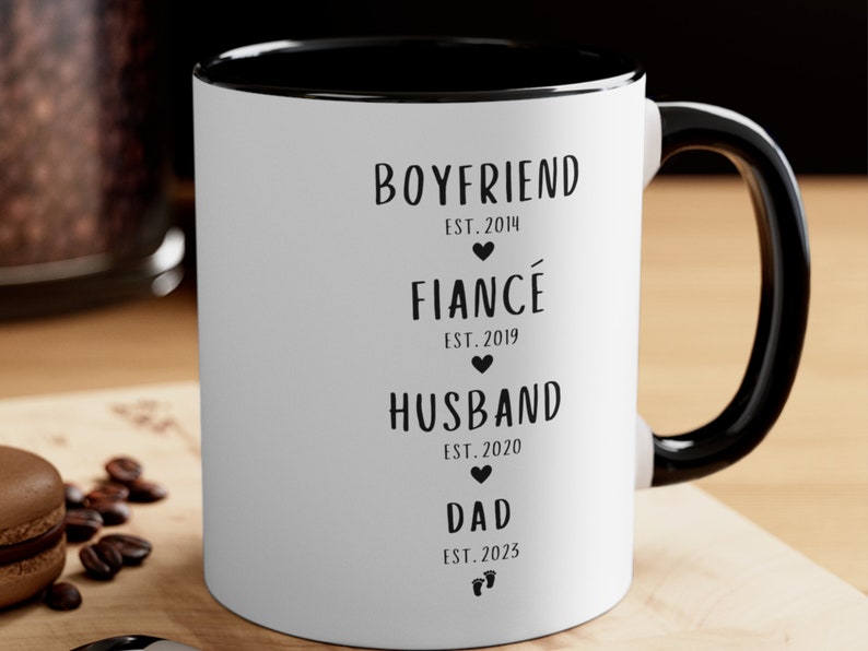 K&ouml;nnte beinhalten: Wei&szlig;e Keramiktasse mit schwarzem Rand und Henkel. Die Tasse hat schwarzen Text mit der Aufschrift "BOYFRIEND EST. 2014", "FIANC&Eacute; EST. 2019", "HUSBAND EST. 2020" und "DAD EST. 2023".
