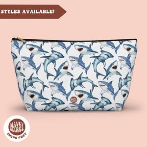 May include: White zippered pouch with a repeating pattern of blue watercolor sharks. The pouch has a black zipper and a gold zipper pull. The text "Wavey Wares Ships Fast" is printed on a brown circle.