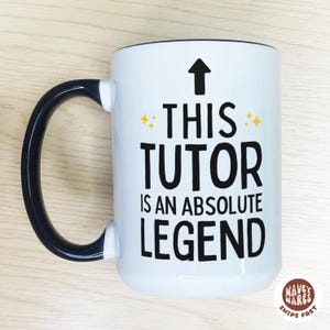 Op de afbeelding: Witte keramische mok met een zwarte handgreep en rand. De mok heeft de tekst "THIS TUTOR IS AN ABSOLUTE LEGEND" in zwart, met een pijl en sterretjes. De mok heeft een glanzende afwerking.