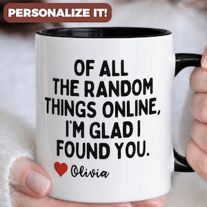 May include: White ceramic mug with a black interior and handle. The mug features the text "OF ALL THE RANDOM THINGS ONLINE, I'M GLAD I FOUND YOU." with a red heart and the name "Olivia". The mug is held by a person.
