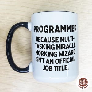 Pode incluir: Caneca de cerâmica branca com alça e borda pretas. A caneca apresenta o texto "PROGRAMMER BECAUSE MULTI-TASKING MIRACLE WORKING WIZARD ISN'T AN OFFICIAL JOB TITLE." A caneca tem o logotipo "WAVEY WARES".