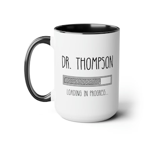 May include: White ceramic mug with a black rim and handle. The mug has a black and white graphic of a progress bar with the text "DR. THOMPSON LOADING IN PROGRESS..."