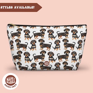 Puede incluir: Bolsa de maquillaje blanca con cremallera con un patrón repetido de perros salchicha negros y tostados. La bolsa tiene una cremallera negra y un tirador de cremallera dorado.