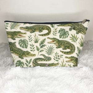 May include: A zippered pouch featuring an off-white background with a repeating pattern of green crocodiles and various tropical leaves. The pouch has a black top edge and a silver-toned metal zipper pull, showcasing a detailed botanical print.