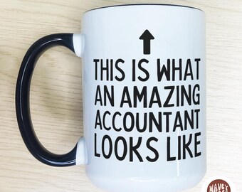 Könnte beinhalten: Weißer Keramikbecher mit schwarzem Henkel und Rand. Der Becher trägt den schwarzen Text „THIS IS WHAT AN AMAZING ACCOUNTANT LOOKS LIKE“ mit einem schwarzen Pfeil nach oben. Der Becher hat ein glänzendes Finish.