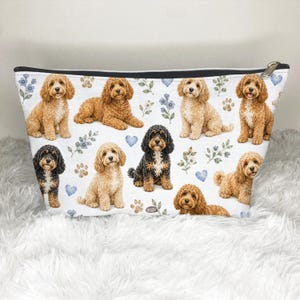 Könnte beinhalten: Ein weißes Reißverschlusstäschchen mit einem sich wiederholenden Muster aus goldbraunen sowie schwarz-lohfarbenen Doodle-Hunden in verschiedenen Posen, durchsetzt mit kleinen blauen Herzen, Pfotenabdrücken und zarten blauen Blumenzweigen.