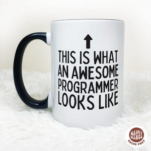 May include: White ceramic mug with a black handle and rim. The mug features the text "THIS IS WHAT AN AWESOME PROGRAMMER LOOKS LIKE" in black, with an upward-pointing arrow above the text.
