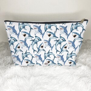 May include: White cosmetic bag with a repeating pattern of blue watercolor sharks. The bag has a black zipper and a gold-colored zipper pull. The bag is sitting on a white, fluffy surface.