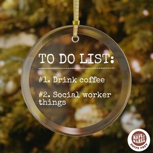 May include: Clear, round Christmas ornament with gold ribbon. The ornament has the text "TO DO LIST: #1. Drink coffee #2. Social worker things". The background is a blurred Christmas tree.