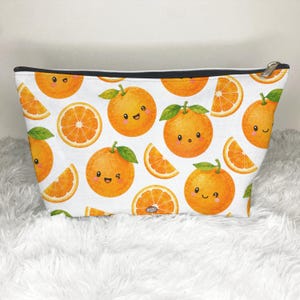 May include: A white zippered pouch featuring a repeating pattern of bright orange citrus fruits. The design includes whole oranges with smiling faces and green leaves, alongside sliced orange segments, all rendered in vibrant shades of orange.