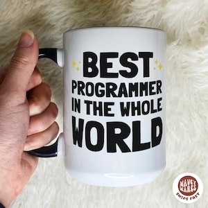 May include: White ceramic mug with a black handle and rim. The mug features the text "BEST PROGRAMMER IN THE WHOLE WORLD" in bold black letters with yellow star accents. The mug is held by a hand.