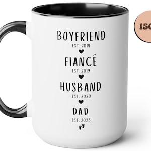K&ouml;nnte beinhalten: Eine wei&szlig;e Keramiktasse mit schwarzem Rand und Henkel. Die Tasse hat schwarzen Text, der "Boyfriend Est. 2014", "Fianc&eacute; Est. 2019", "Husband Est. 2020" und "Dad Est. 2023" lautet. Die Tasse hat ein Fassungsverm&ouml;gen von 440 ml.