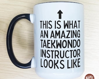 May include: White ceramic mug with a black handle and rim. The mug features the text "THIS IS WHAT AN AMAZING TAEKWONDO INSTRUCTOR LOOKS LIKE" in black, with an arrow pointing upwards. The mug has a glossy finish.