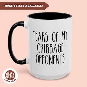 Può includere: Una tazza da caffè in ceramica bianca con manico e bordo neri. La tazza ha la scritta "Tears of my Cribbage Opponents" stampata in nero.