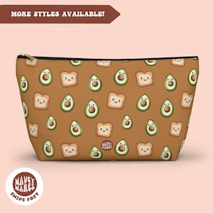 Può includere: Una pochette con cerniera marrone con un motivo ripetuto di metà avocado e fette di pane tostato con facce felici. La pochette ha una cerniera nera e un bordo nero.