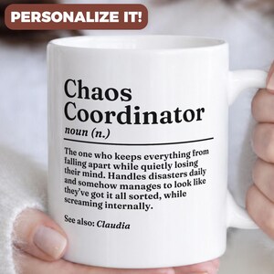 May include: White ceramic mug with the text "Chaos Coordinator" in black. The mug includes a definition of the term and the words "See also: Claudia". The mug has a handle and is held by a person.