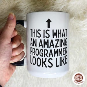 May include: White ceramic mug with a black handle and the text "THIS IS WHAT AN AMAZING PROGRAMMER LOOKS LIKE" in black. An arrow points upwards. The mug is held by a hand.