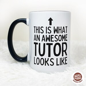 May include: White ceramic mug with a black handle and rim. The mug features the text "THIS IS WHAT AN AWESOME TUTOR LOOKS LIKE" in black, with an upward-pointing arrow above the text.